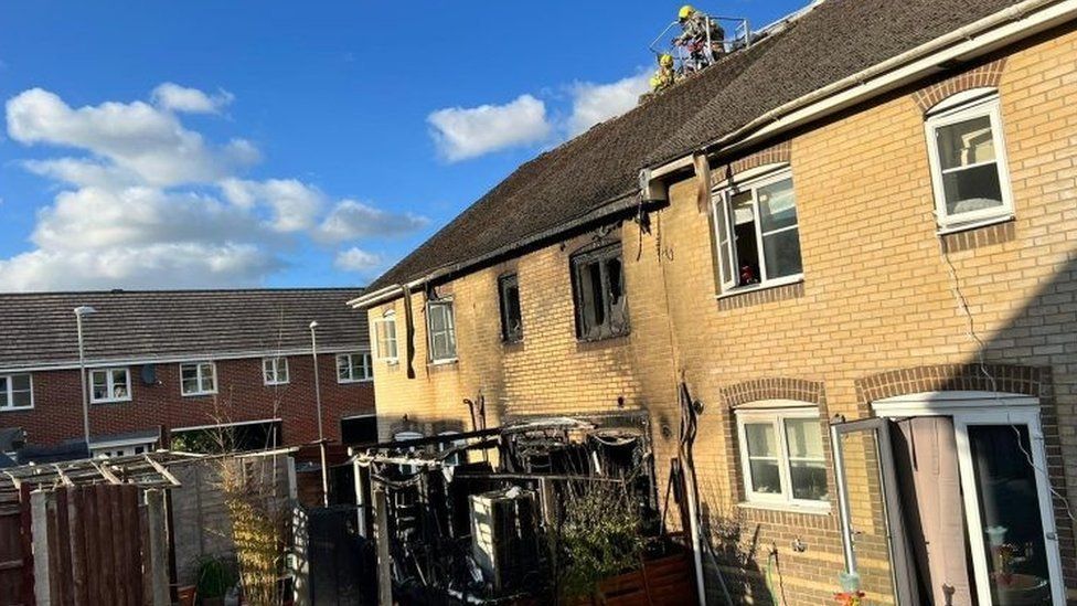 Firefighters on the roof of the pale brick mid-terraced house with the burnt-out shell of a conservatory in the garden and blackened first-floor windows from the blaze