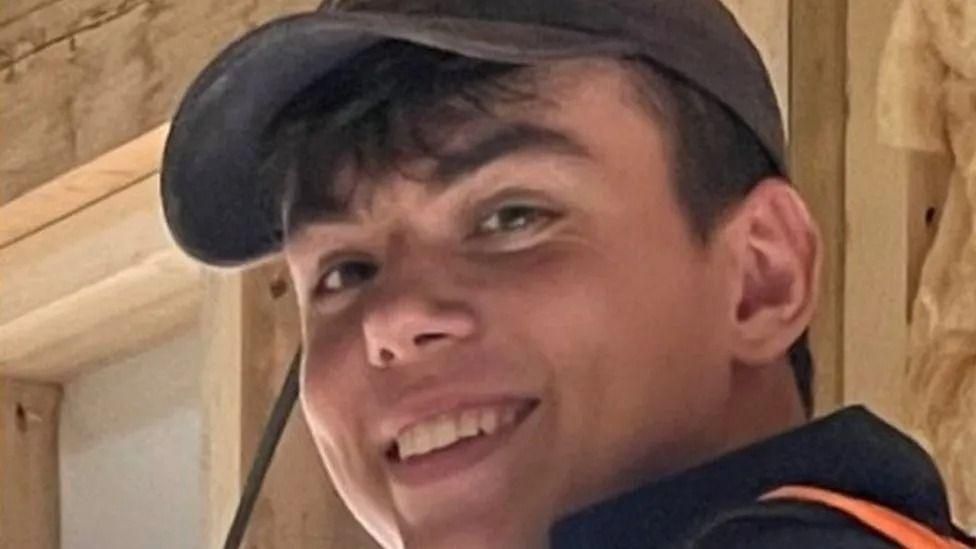 A slightly blurry photo of a young man with short dark hair wearing a black baseball cap and smiling at the camera.