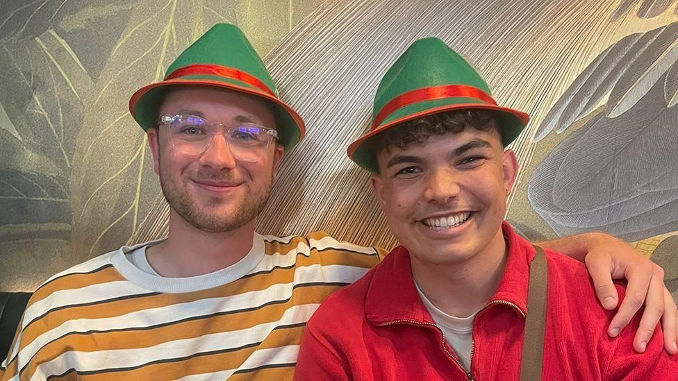 One man in yellow and white stripy t-shirt wearing a red and green hat, with his arm around another man who has a red top on and the same red and green hat