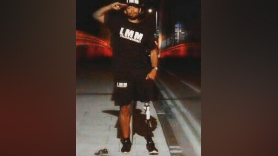 Image of Jade Anthony Barnett, a man with a prosthetic leg making a salute while dressed in a black top, shorts and baseball cap