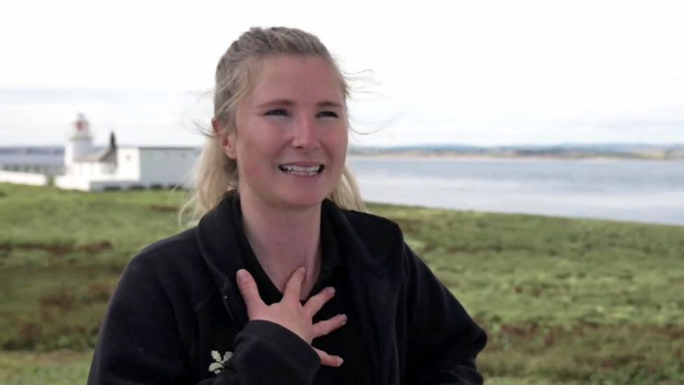 Sophia Jackson, who has blonde hair, holds her hand to her chest looking happy. A white lighthouse is in background. 