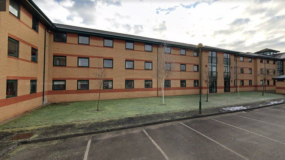 A four-storey block of flats, with a small grassed area in front of the building and an empty car park alongside 