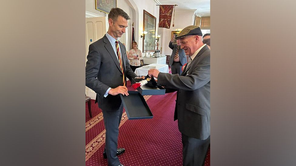 Major General Jon Swift, dressed in a dark grey suit, presents Fusilier Steve Close, also wearing a grey suit, with a presentation box containing his beret and hackle, made up of red and white feathers. Several people are smiling in the background