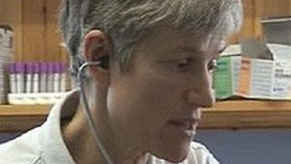 Dr Sarah Myhill close up of her face... she has a stethoscope in her ears and medicines on a shelf behind her
