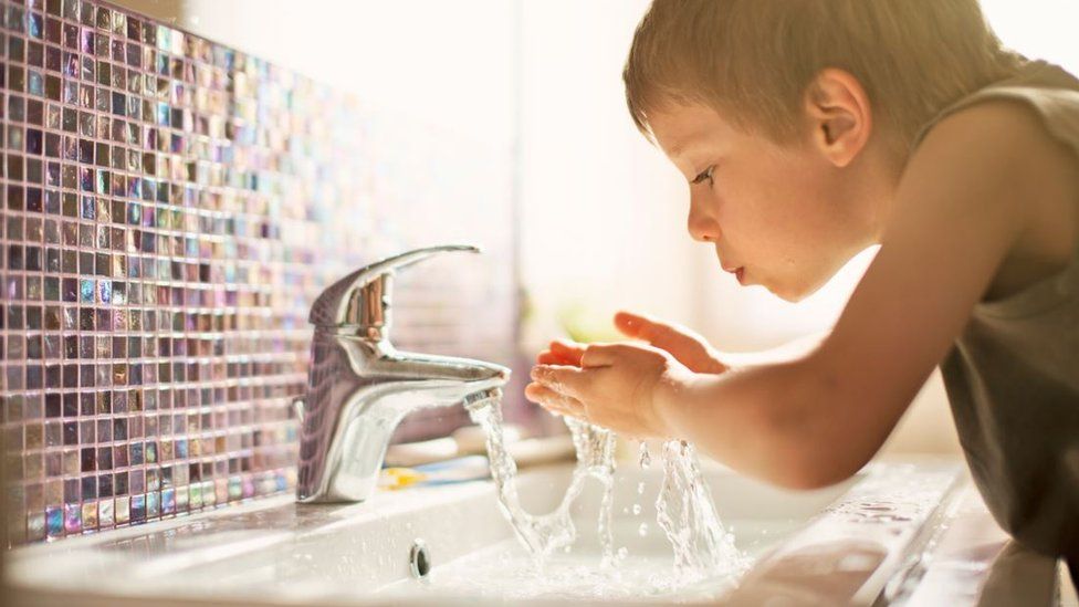 A little boy splashing his face with water from a mixer tap