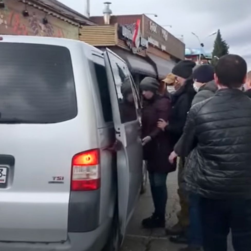 A silver minibus with an open door and people standing next to it