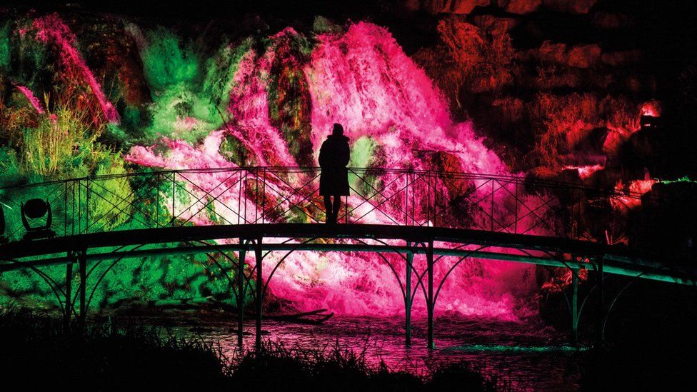 Silhouette of a person standing on a bridge against a pink illuminated waterfall