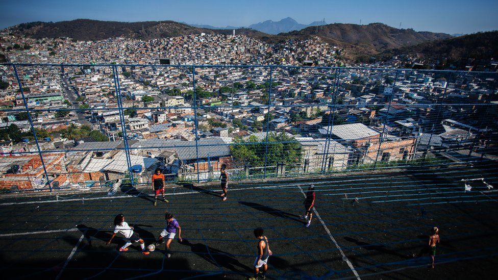 A wide shot of the favela beyond the chain-link fence of the training field
