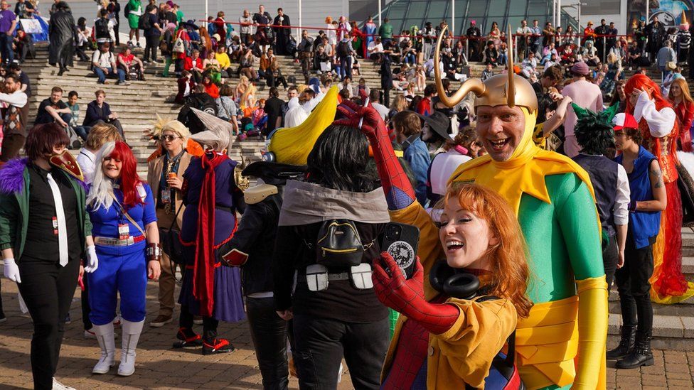 A woman dressed as Spider Man takes a selfie of herself and a man dressed as Loki in front of crowds