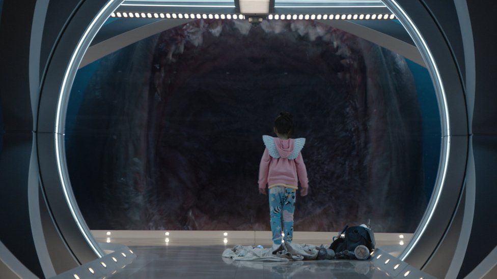A small child stands in front of a large shark with its jaws open
