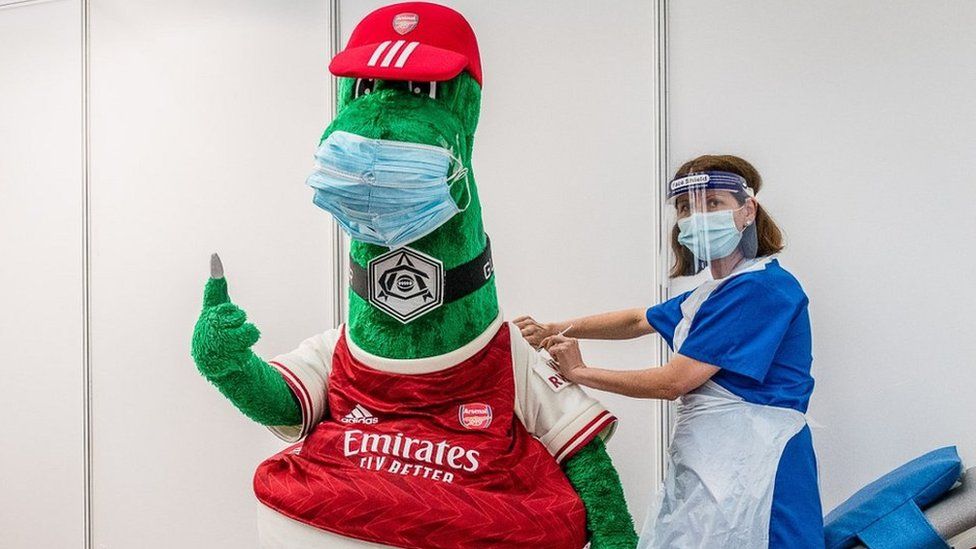 Arsenal mascot Gunnersaurus (left) gets his Covid vaccination from an NHS frontline worker