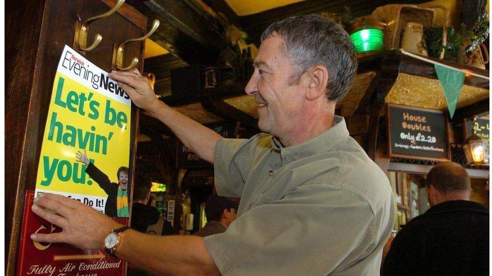 Mr Brown putting up a poster in one of his pubs. He is wearing a grey/green shirt and smiling.