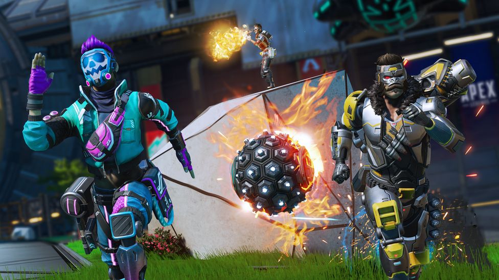 Two characters in futuristic armour sprint towards the viewer. The character on the left wears a sky blue suit of armour with a skull-face mask and bright purple trim. The character on the right wears a silver suit of armour with yellow trim and a mask styled on a gorilla's face. They're fleeing from a large, spiked metal ball with flames emanating from it. Behind them, stood atop a piece of wreckage, a third character fires at an unseen enemy - a volley of flame emanating from their weapon.