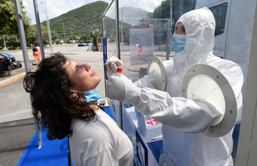 A person in protective clothing swabs a woman's nose, with a transparent barrier diving them
