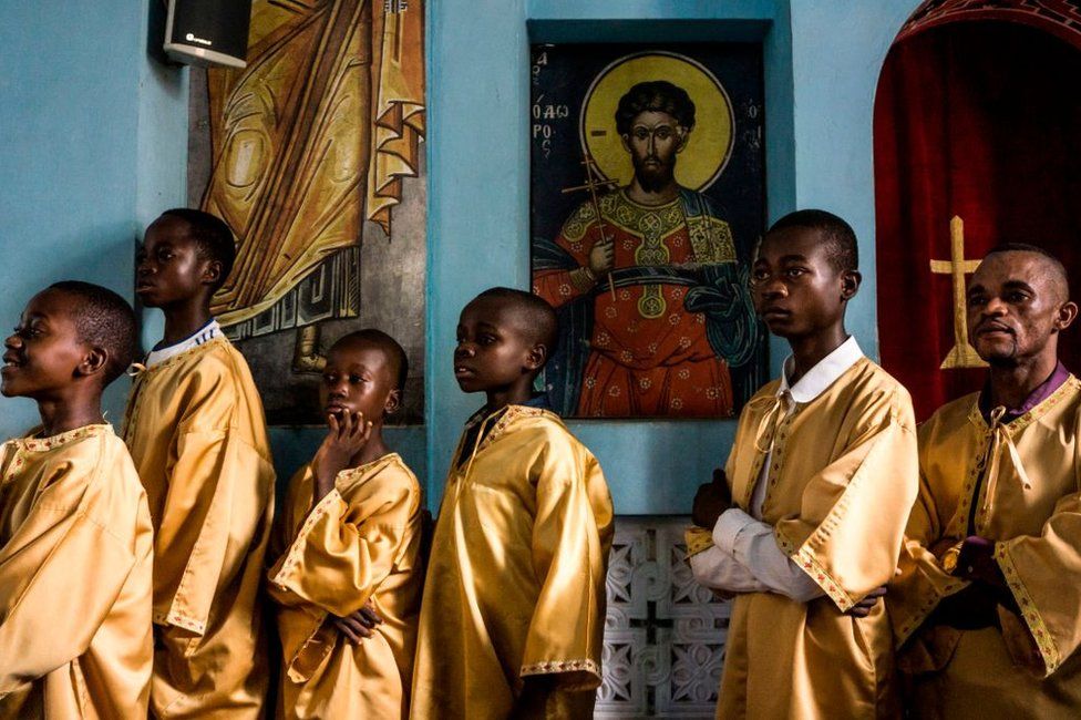 Boys and men dressed in golden robes stand next to ornate religious paintings inside the church