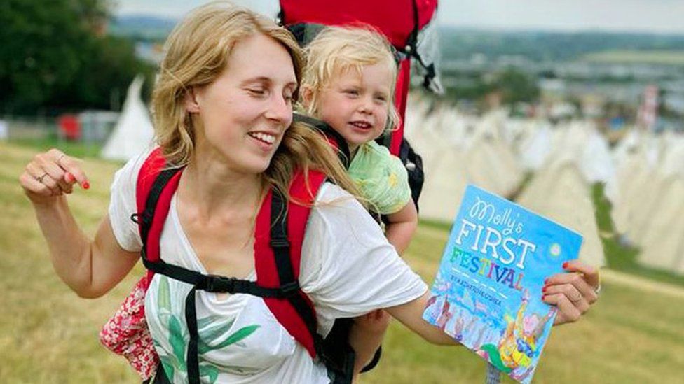 A young blonde haired woman with a young toddler in a baby carrier on her back