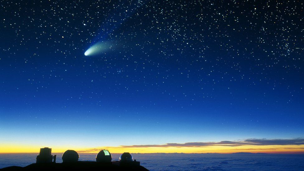 A row of telescopes above the cloud tops, with a comet streaking across the sky and stars visible in the background