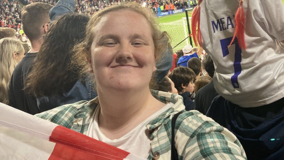 Khya Gott at a football match. Khya is a white woman with curly blonde hair tied back. She smiles at the camera, her eyes squeezed shut and holds up an England flag. She wears a green chequered shirt over a white T-shirt and behind her fans are cheering. The pitch and a goal can just be made out.