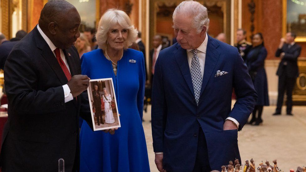 Г-н Рамафоса держит фотографию королевы Елизаветы II с Нельсоном Манделой, когда он разговаривает с Камиллой и королем Карлом III.