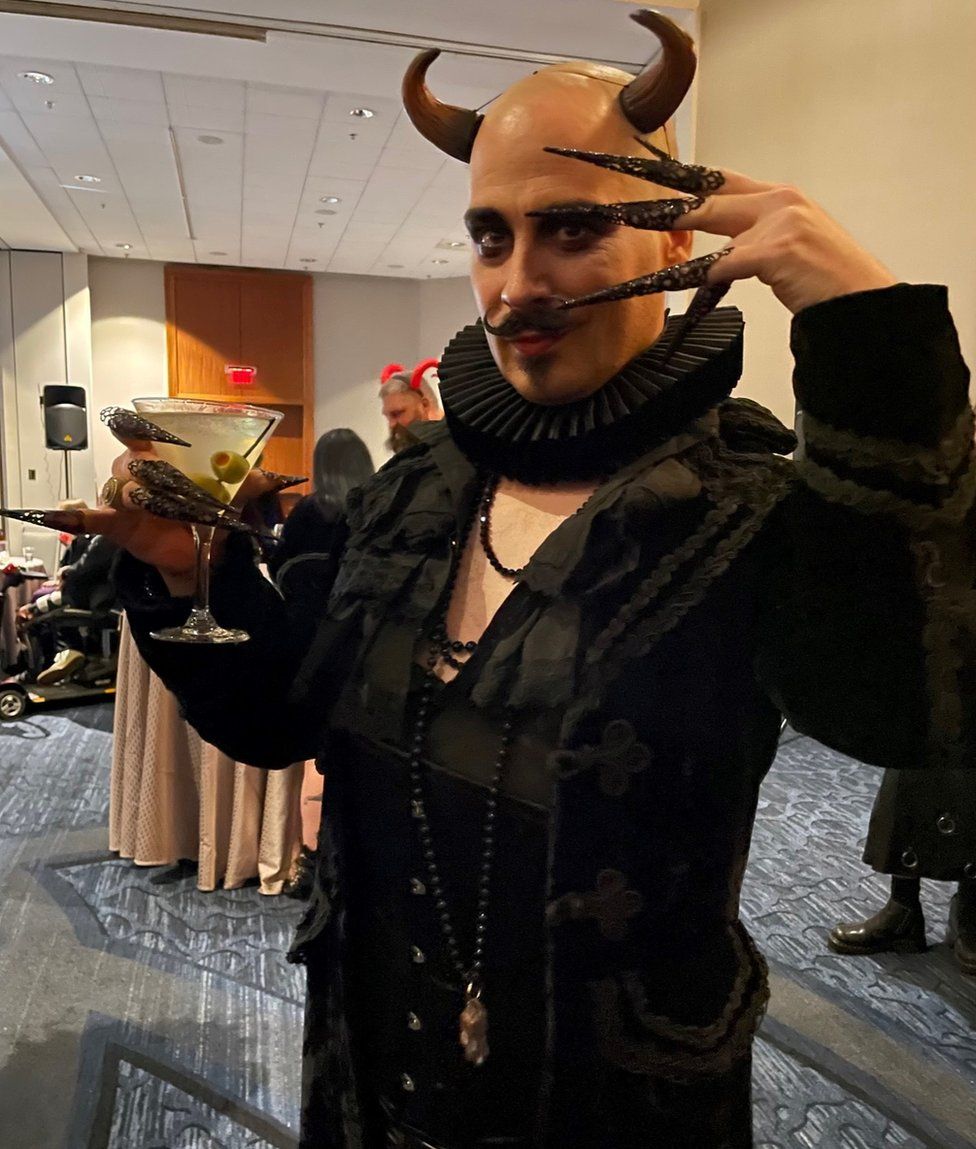 A man wearing horns, a black ruff and elaborate pointed fake nails poses with a martini