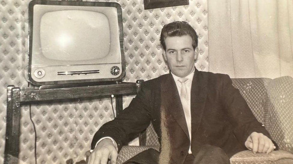 A black and white photo of Laurie Cornish. He is wearing a suit, shirt and tie and sitting in an armchair. Next to him is a television on a stand. 