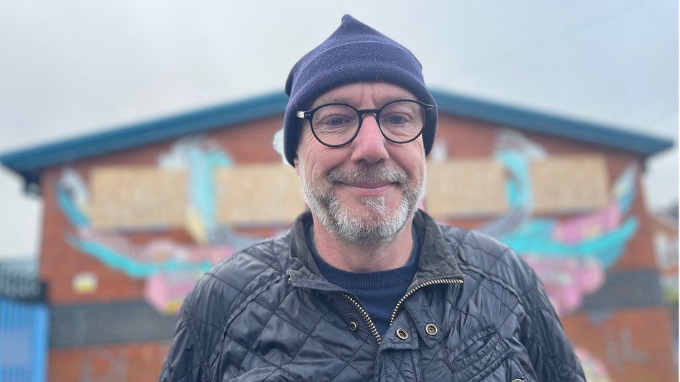 a white man in a hat with glasses outside a derelict building