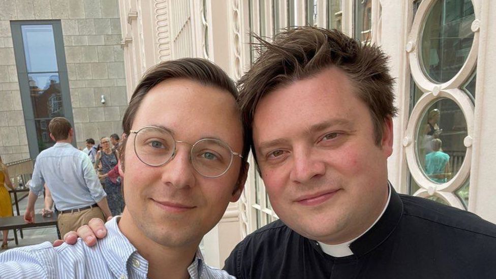 Charlie Bell, a young man in a priest's dog collar poses for a picture with his partner Piotr, who wears glasses.