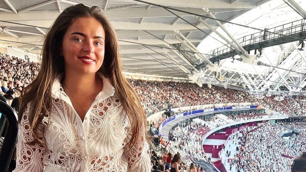 Liliana Cooper - pictured in her restricted view seat when she saw The Weeknd in London. Liliana is a young white woman with a tan - she has with green eyes and long brown hair and is smiling at the camera. She is wearing a white lacy top and is pictured standing in front of thousands of other fans in the stadium.