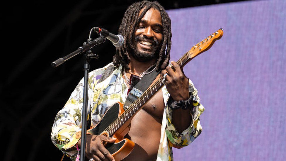 Hak Baker performing on stage. He is smiling and holding a black guitar in front of a mic. He has shoulder length dreadlocks and is wearing a patterned shirt that is unbuttoned all the way down