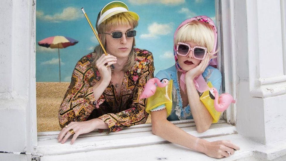 A couple dressed in colourful summer clothes look out of a window on to a beach