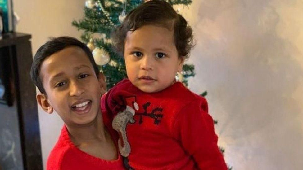 A picture of two boys in front of a Christmas tree
The older boy is smiling, with black hair, wearing a red t shirt. He is holding the younger boy, with brown hair who is wearing a ted top with a reindeer on it