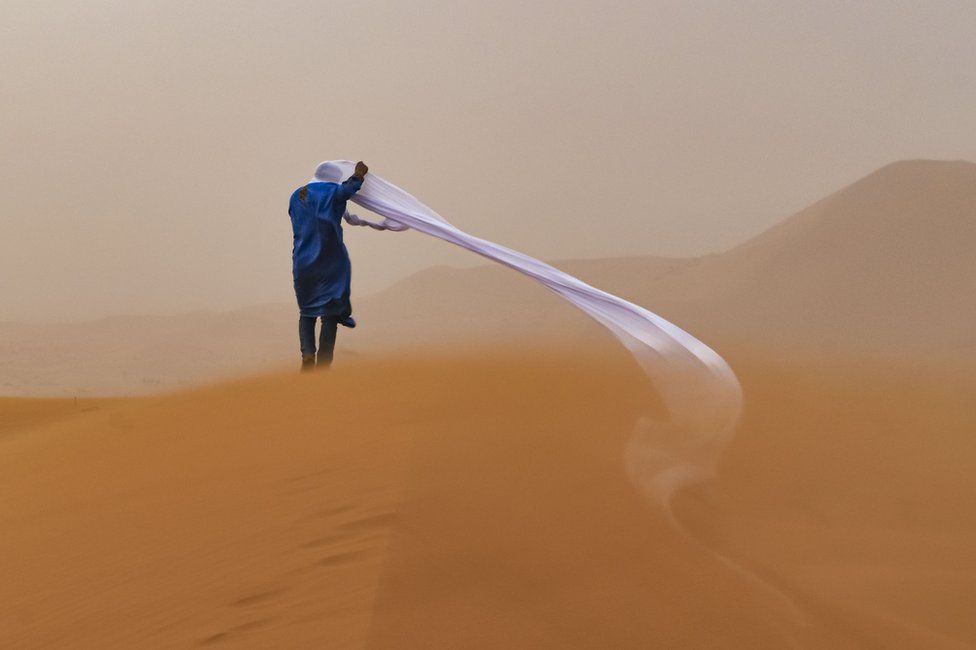 A man struggles with wind blowing at cloth around his head whilst standing in the Sahara Desert