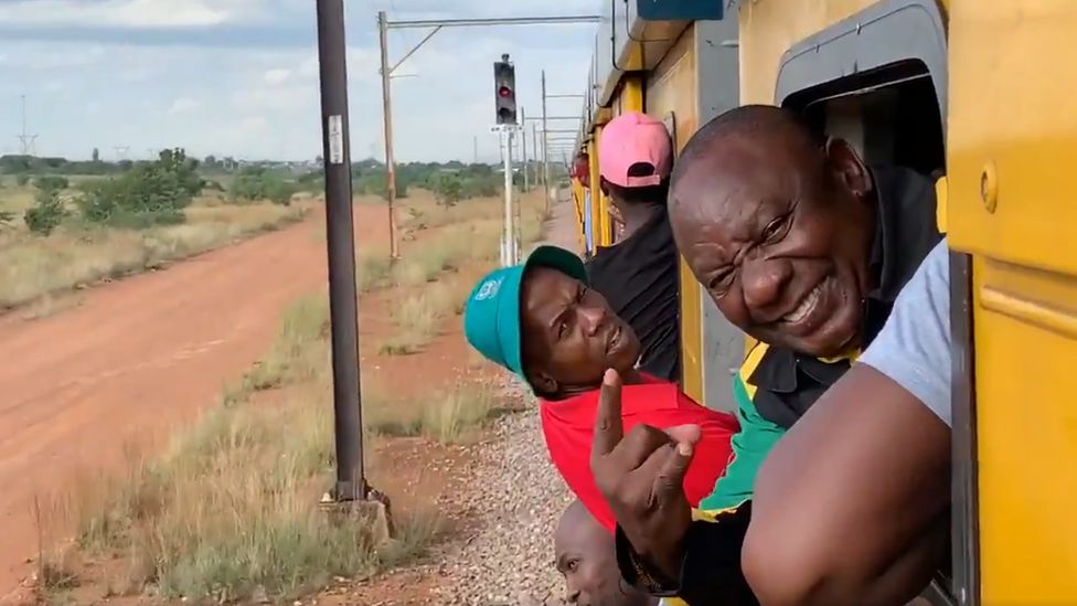 President Cyril Ramaphosa is seen smiling, gesturing and sticking his head out of a train window