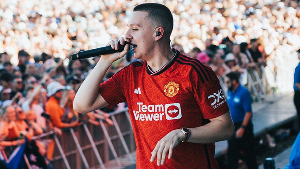 Aitch wearing the new Manchester United shirt, which is red and has the team logo and Adidas sponsor, along with the name of their Team Viewer sponsor in the middle of the shirt. He is wearing white trousers, and is holding a microphone, and has a watch on his left hand. The background has thousands of people watching Aitch perform.