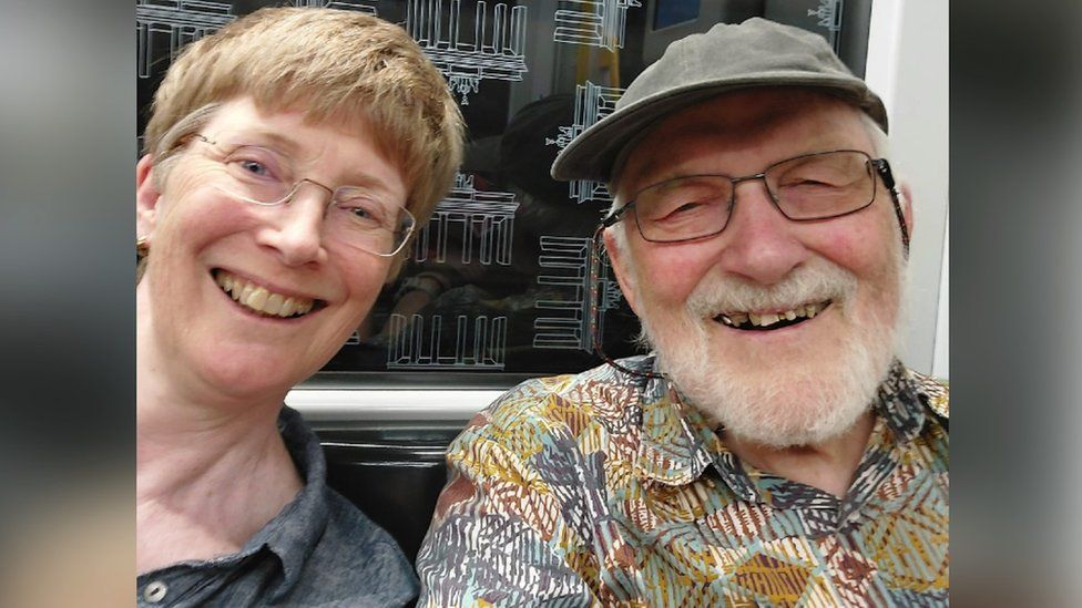 Roger Sturge pictured with his daughter, Dr Kate Sturge. They are sat next to each other travelling on the U-Bahn in Berlin. Both are looking at the camera and smiling.