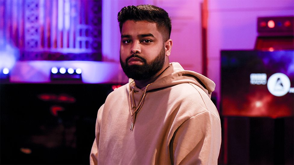 Ezu, a bearded Asian man, wearing a light brown hoody. Behind him are screens and a wall which is illuminating purple from lights. The screen is slightly blurred but has a logo which reads BBC Asian Network.