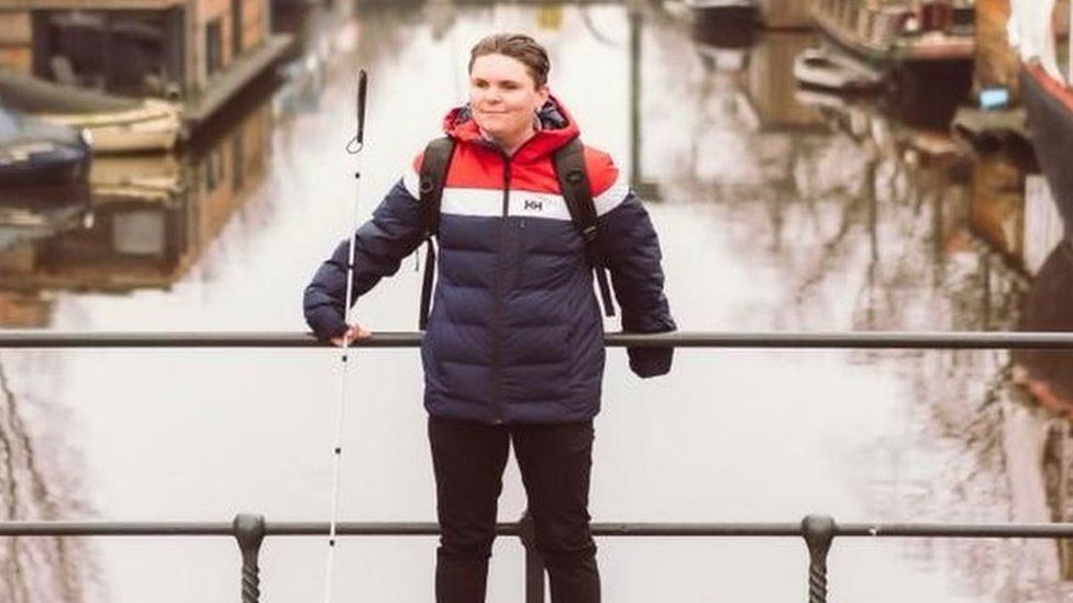 Connor Scott-Gardner stood wearing black boots and black trousers with a navy, white and red coat and his white cane leaning against a bridge going over a canal