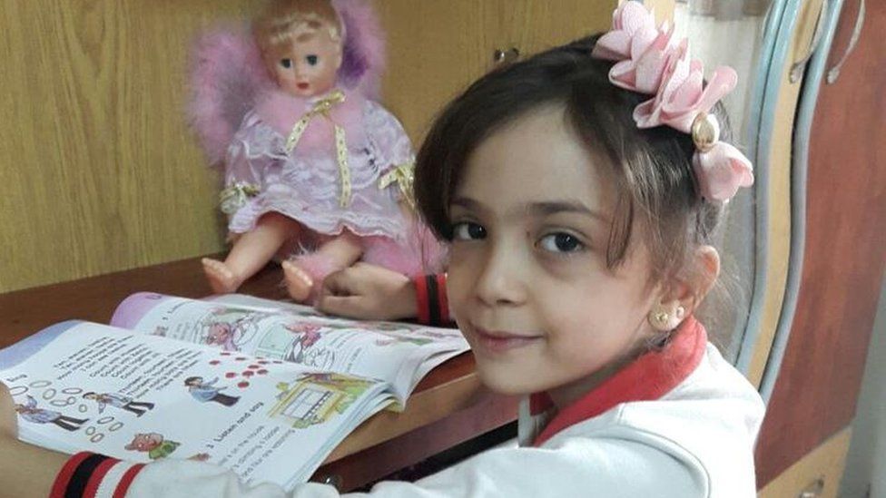 Bana Alabed, seated at a desk with a book and doll, from her Twitter account