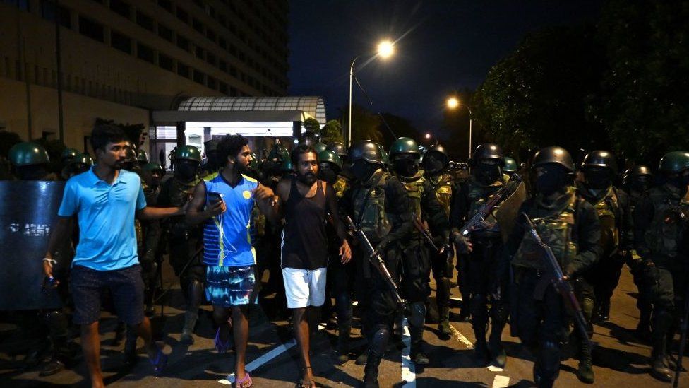 Members of Sri Lankan security forces walk beside people at the site of protest camp