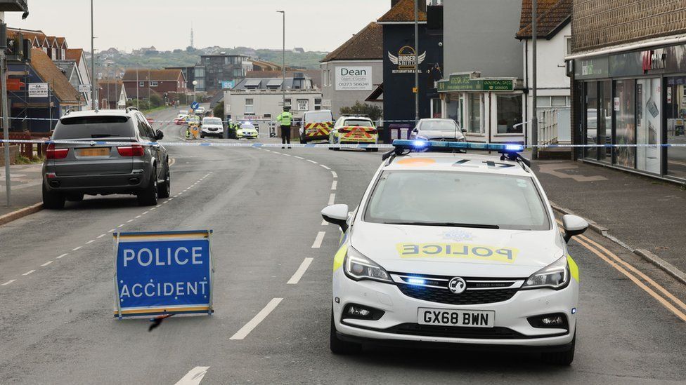 A police car at the scene of the crash on a road that has been taped off with an officer standing guard