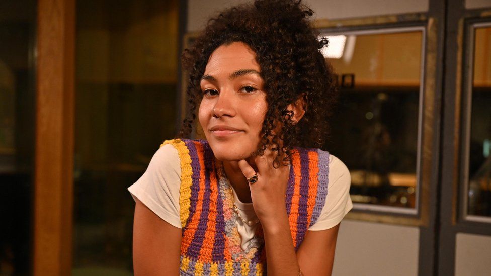 Olivia Dean pictured at the BBC's Maida Vale studio while performing for Radio 1's Future Sound session in March 2023. The 24-year-old singer wears a colourful crochet vest over a white T-shirt and rests her chin on her right hand while leaning on a surface. She has brown eyes and wears her dark curly hair tired back.