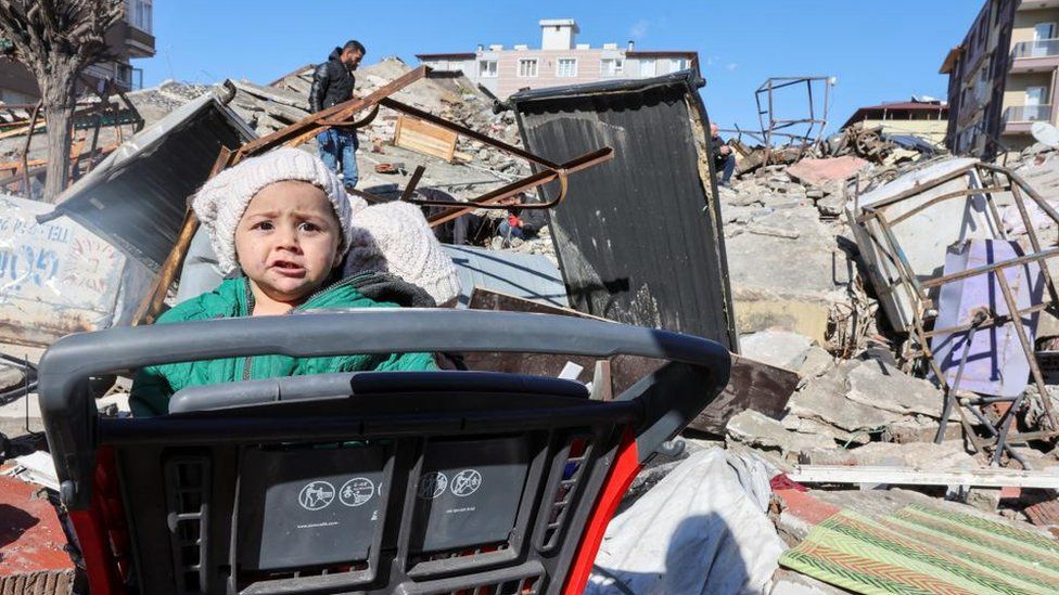 A child sits in a shopping cart at the site of a collapsed building, in the aftermath of a deadly earthquake in Hatay, Turkey, February 8, 2023