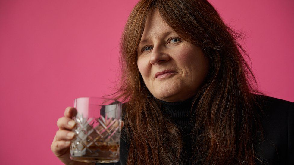 Joyce Greenaway raising a glass of whiskey against a pink backdrop