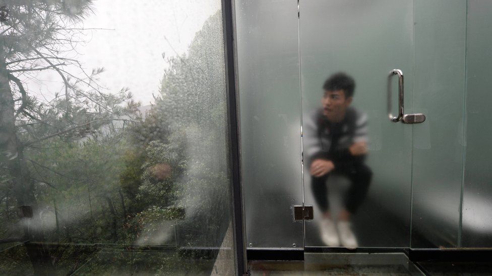 Man sits on toilet, behind lightly frosted glass door, looking out at the forest outside. Changsha, China, 29 September 2016.
