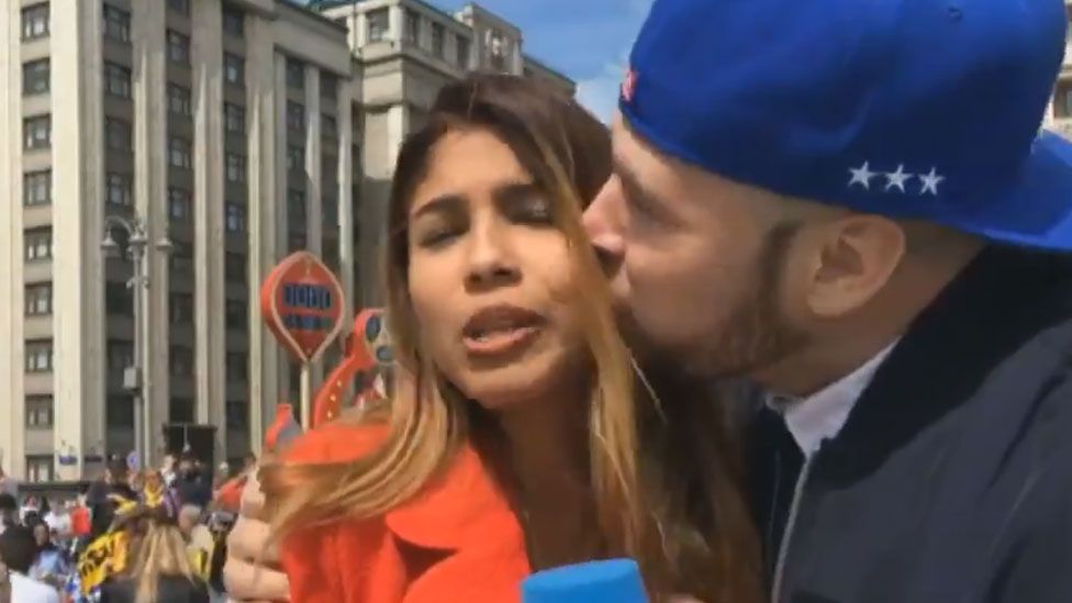 A female reporter is kissed, by a fan, clearly looking uncomfortable, holding a DW mic