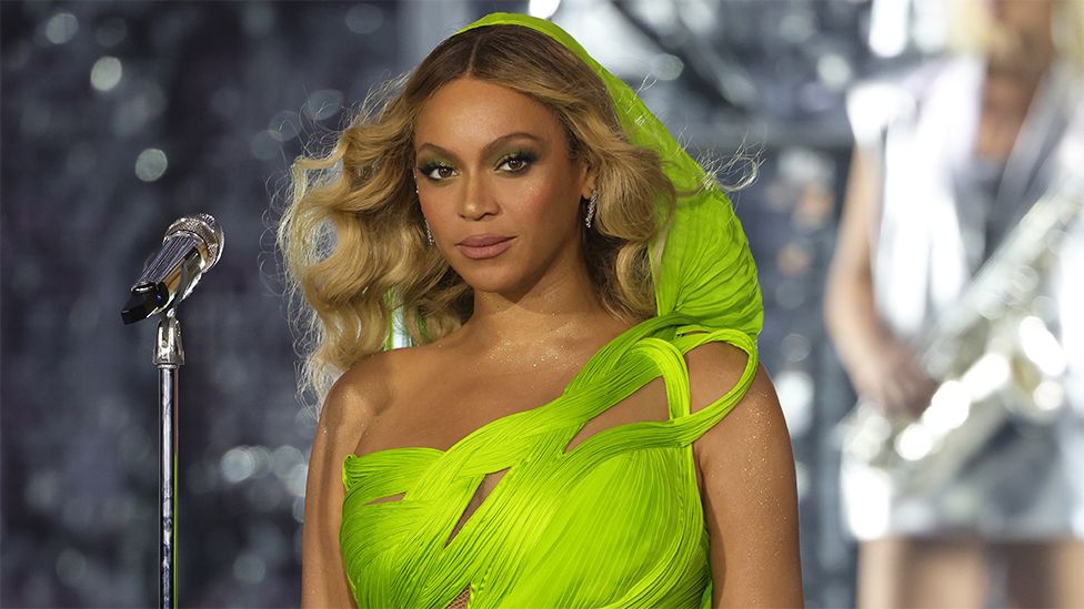 Beyoncé wearing Gaurav's design on her Renaissance world tour. The dress is neon green and has a flowing hood and a knotted design on one shoulder. Beyoncé matches the dress with green eye shadow and has curled ombre blonde hair. She stands in front of a chrome microphone and looks at the camera