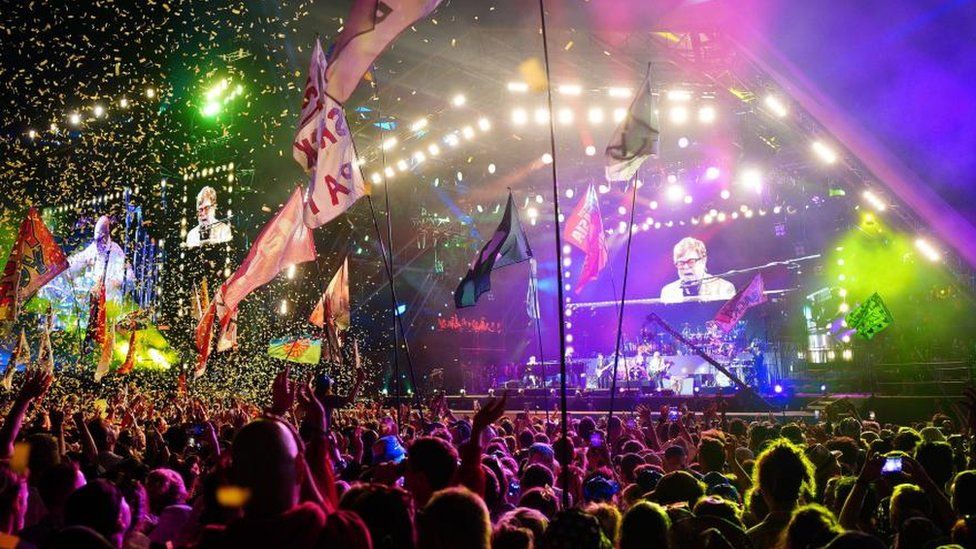 Elton John performing on stage at Glastonbury. The photo is taken from the perspective of the crowd so you can see the backs of a lot of heads in the crowd and lots of flags and confetti in the air. You can make out Elton John signing at his piano on the big screen at the back of the stage.