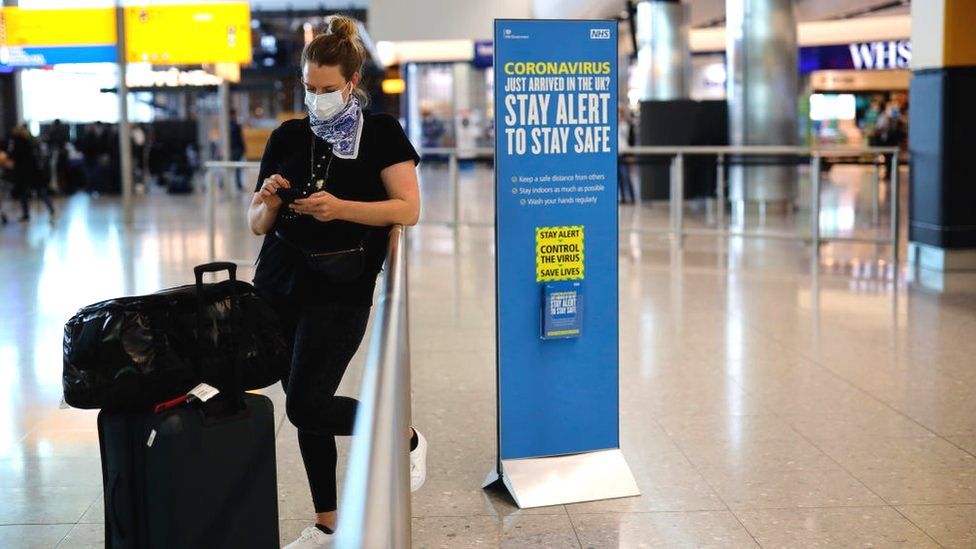 A woman wearing a mask in the departures hall at Heathrow Airport, standing next to a sign