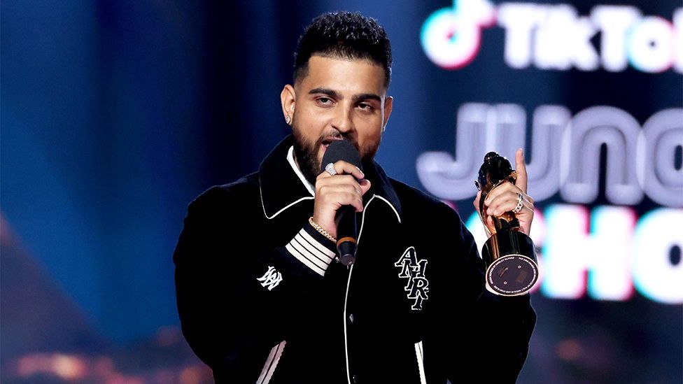 A man holding a black microphone and trophy in his hands, wearing a black jacket.