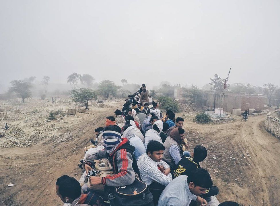 The top of a train is crowded with men as it speeds along dirt roads.